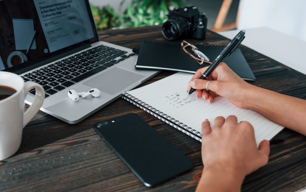 Person writing in a notebook beside a laptop and phone.
